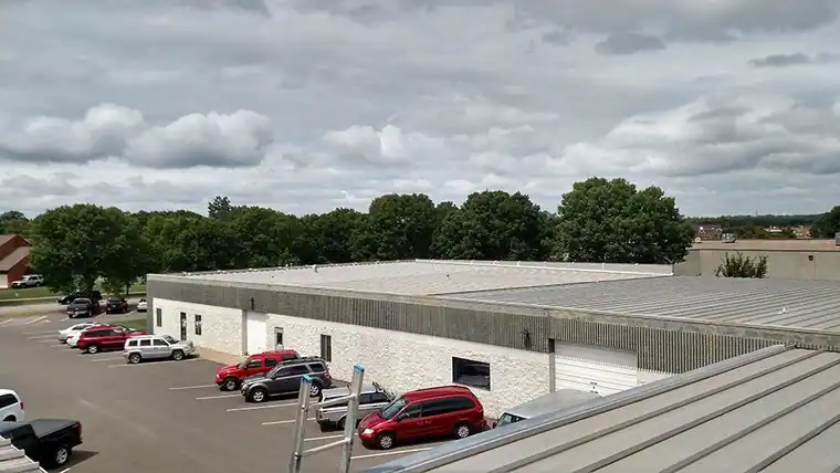 Long white industrial building with a low-slope metal roof beside a parking lot